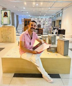 Imagem do idealizador da exposição, Diego Rimaos, servidor do APESP. Ele está sentado em uma estrutura de madeira, segurando uma pasta com papéis dentro. Ao fundo, a estrutura da exposição "Eu amanuense que escrevi...".