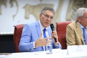 Homem fala ao microfone de uma bancada vestindo um terno azul