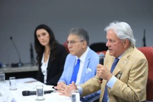 Três pessoas numa bancada, sendo dois homens e uma mulher. O homem que veste terno claro fala ao microfone