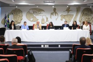 A foto mostra uma mesa de autoridades em um auditório. Seis pessoas estão sentadas lado a lado atrás de uma mesa coberta com toalha branca, enquanto um público acompanha de poltronas vermelhas. No centro, um dos participantes fala ao microfone. Ao fundo, há um painel artístico em relevo com figuras humanas e símbolos, além de bandeiras posicionadas à esquerda. O ambiente é formal e indica a realização de um evento institucional ou conferência.