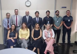 "Uma foto de grupo de dez pessoas — sete homens em pé na fileira de trás e quatro mulheres sentadas na fileira da frente — em uma sala de reunião ou escritório. A maioria dos homens veste camisas sociais e ternos ou gravatas, enquanto as mulheres vestem trajes sociais de trabalho. O ambiente é formal, com paredes claras, um relógio na parede superior central e uma porta à direita."