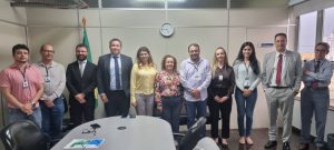 "Uma foto de grupo de onze pessoas — sete homens e quatro mulheres — em pé atrás de uma mesa de reunião oval em um escritório. A maioria veste trajes casuais de negócios ou sociais. Da esquerda para a direita, os homens vestem uma camisa rosa, uma camisa cinza-claro, um terno preto, um terno azul-escuro, uma camisa lilás e cinza-claro. Uma mulher no centro veste uma blusa floral colorida. A mesa de reunião é grande e tem alguns objetos sobre ela. Ao fundo, há uma bandeira do Brasil, um relógio de parede e uma janela com vista para prédios da cidade."