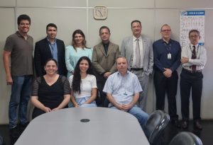 "Uma foto de grupo de dez pessoas — sete homens e três mulheres — em uma sala de reunião ou escritório. Sete pessoas estão em pé na fileira de trás, e três estão sentadas na fileira da frente, atrás de uma mesa oval. A maioria veste trajes sociais ou casuais de negócios, como camisas polo, camisas sociais, blazers e ternos. Ao fundo, há paredes claras, um relógio de parede no centro e um pôster informativo à direita."