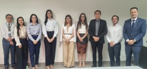Um grupo de nove pessoas, incluindo três homens de terno ou camisa social e seis mulheres em trajes de negócios e elegantes, posando lado a lado em um escritório.