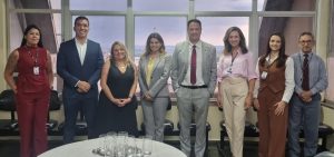 "Uma foto de grupo com oito pessoas — cinco mulheres e três homens — em pé em uma sala com grandes janelas de onde se vê a paisagem urbana. Todos estão vestidos com trajes sociais ou casuais de negócios. Da esquerda para a direita: uma mulher de conjunto bordô; um homem de terno azul-marinho; uma mulher de vestido preto; uma mulher de terno cinza com blusa amarela; um homem de terno cinza com gravata bordô; uma mulher de camisa rosa e calça bege-claro; uma mulher de colete marrom e calça marrom; e um homem de terno cinza com gravata bordô. Na frente do grupo, há uma mesa com uma bandeja de metal com vários copos vazios."