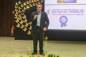 Foto de um homem de pele clara e cabelos grisalhos. Ele está de pé e falando ao microfone. O homem usa um terno preto e uma camisa azul claro.