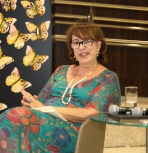 Foto de uma mulher de pele clara e cabelos castanhos. Ela está sentada em uma cadeira bege e usa um vestido azul com estampa de flores e óculos de grau.