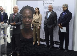 Homem de terno fala ao microfone. Atrás pessoas assistem à fala