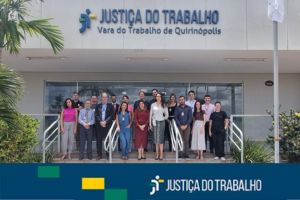A imagem mostra um grupo de pessoas em frente a um prédio com a inscrição "JUSTIÇA DO TRABALHO - Vara do Trabalho de Quirinópolis". As pessoas, vestindo trajes sociais ou casuais, estão posando para a foto na escadaria de entrada do edifício. A parte inferior da imagem tem uma faixa azul escura e verde-amarela com o logo da Justiça do Trabalho.