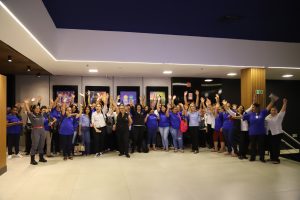 Um grande grupo de mulheres, muitas vestindo camisetas azuis ou uniformes, de pé e acenando com os braços para cima em um saguão de cinema ou evento.