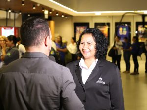 Uma mulher sorridente em um blazer preto sendo entrevistada por um homem (visível de costas) em um saguão movimentado com muitas pessoas e câmeras de vídeo.