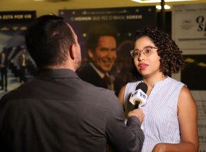 Uma mulher com cabelo cacheado e óculos, usando uma camisa listrada, sendo entrevistada por um homem (visível de costas) que segura um microfone com o logotipo "TRT-18", com pôsteres de filmes no fundo.