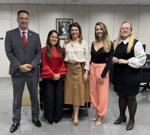 
Um grupo de cinco pessoas, um homem de terno e quatro mulheres em diferentes trajes sociais e de negócios, posando em pé em um escritório.