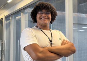Um jovem com cabelo cacheado e óculos, vestindo uma camiseta branca, sorrindo com os braços cruzados em frente a uma vidraça.