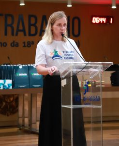 Mulher fala ao microfone em um púlpito do TRT da 18ª Região, vestindo camiseta do programa Trabalho, Justiça e Cidadania.