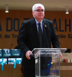 Homem fala ao microfone em um púlpito do TRT da 18ª Região, usando terno escuro e gravata.