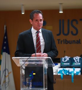 A imagem mostra um homem de terno preto e gravata vinho-escura, com barba, falando em um microfone que segura na mão direita.