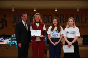 A imagem mostra um grupo de cerca de 14 pessoas, principalmente jovens e mulheres, posando em um auditório. O grupo está em pé em cima de um tapete marrom, e há dois homens de terno nas extremidades e um no centro, atrás. As pessoas na frente seguram sacolas de brindes e uma delas exibe um certificado. O fundo é de madeira, com a frase "JUSTIÇA DO TRABALHO" em destaque na parede e um relógio digital vermelho marcando a hora.