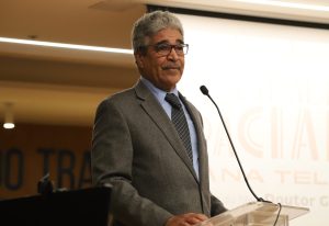 Um homem de meia-idade com cabelos grisalhos, terno e óculos, falando em um pódio de acrílico com um microfone em um auditório.