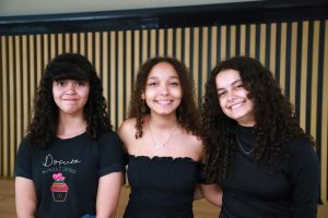 Três jovens posam sorridentes lado a lado em um ambiente interno, diante de uma parede com ripas de madeira.