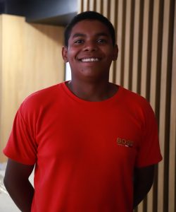 Jovem sorri para a câmera em ambiente interno, vestindo camiseta vermelha diante de uma parede com ripas de madeira.