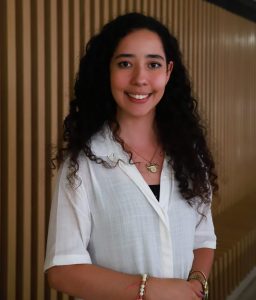 Jovem sorri para a câmera em ambiente interno, vestindo camisa branca e acessórios, diante de uma parede com ripas de madeira.