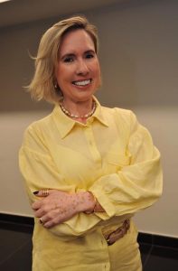 Mulher de pele clara e cabelos loiros. Ela usa blusa amarela e está de braços cruzados e sorrindo