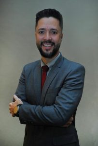 Homem de pele clara e cabelos castanhos. Ele usa terno e está de braços cruzados e sorrindo