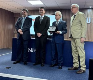 Quatro homens em em um auditório. Um deles segura uma placa de homenagem
