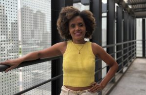 Uma mulher de cabelos cacheados e top amarelo posando em uma passarela moderna, com edifícios altos de vidro no fundo.