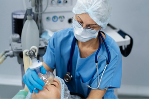 Mulher, profissional da saúde, está de uniforme azul, mascara e touca, colocando uma mascara de oxigenio na paciente deitada numa maca.