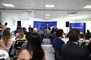 imagem mostra várias pessoas sentadas em mesas com cadeiras e o presidente do TRT ao fundo